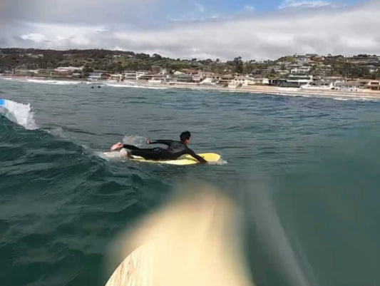 Surfer paddling out on a yellow foam board along the California coastline with a beachside neighborhood in the background
