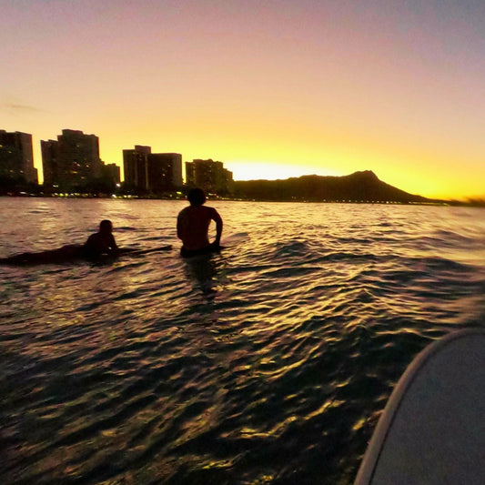 Seas Get Degrees Crew doing a sunrise surf session in Hawaii with sunrise over ocean water with silhouetted swimmers or surfers, distant city buildings, and a mountain skyline.