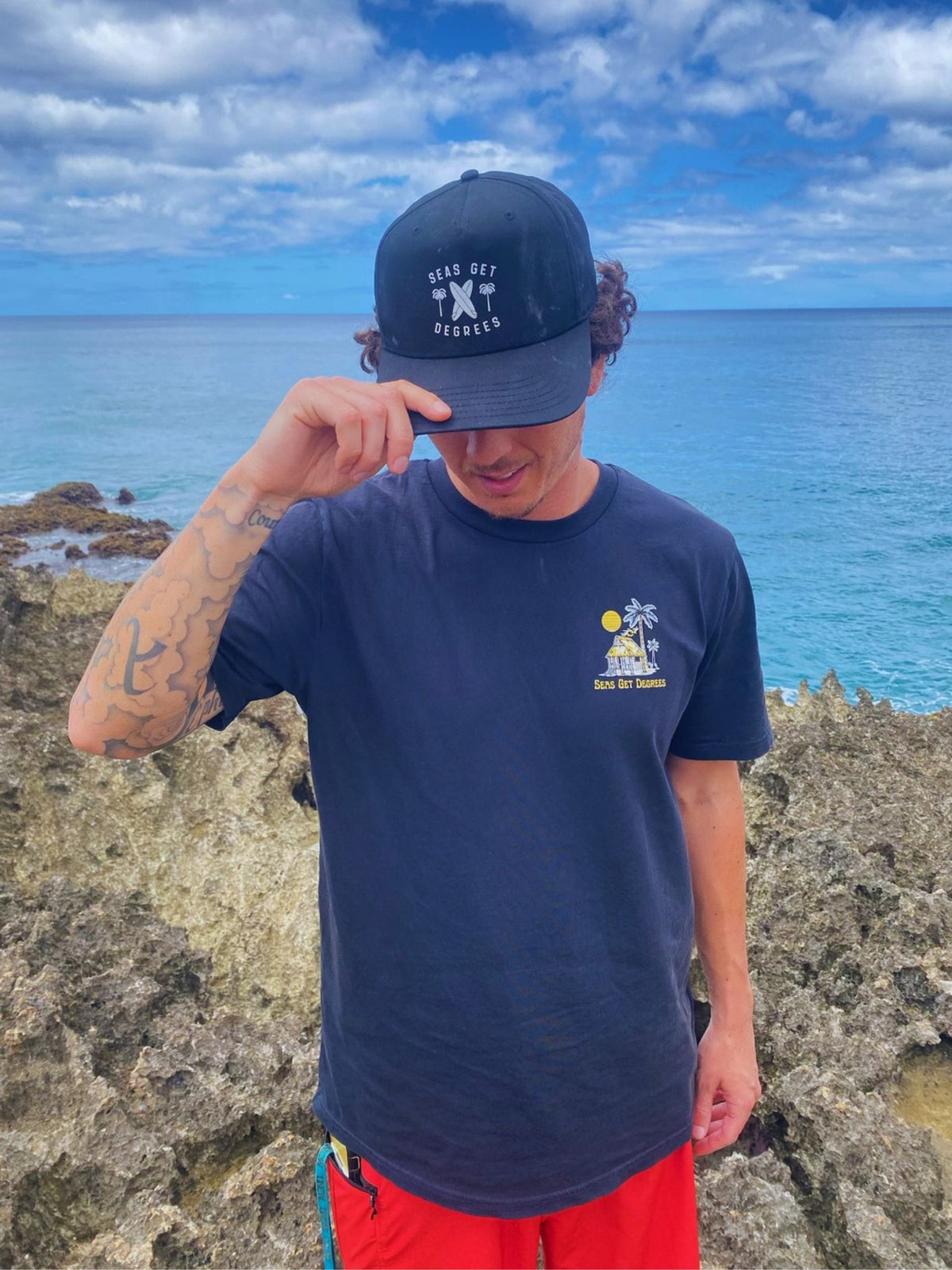 A man stands on a rocky coastline looking out at the ocean, wearing a black "Lost & Found" Seas Get Degrees Tee and with Seas Get Degrees snapback