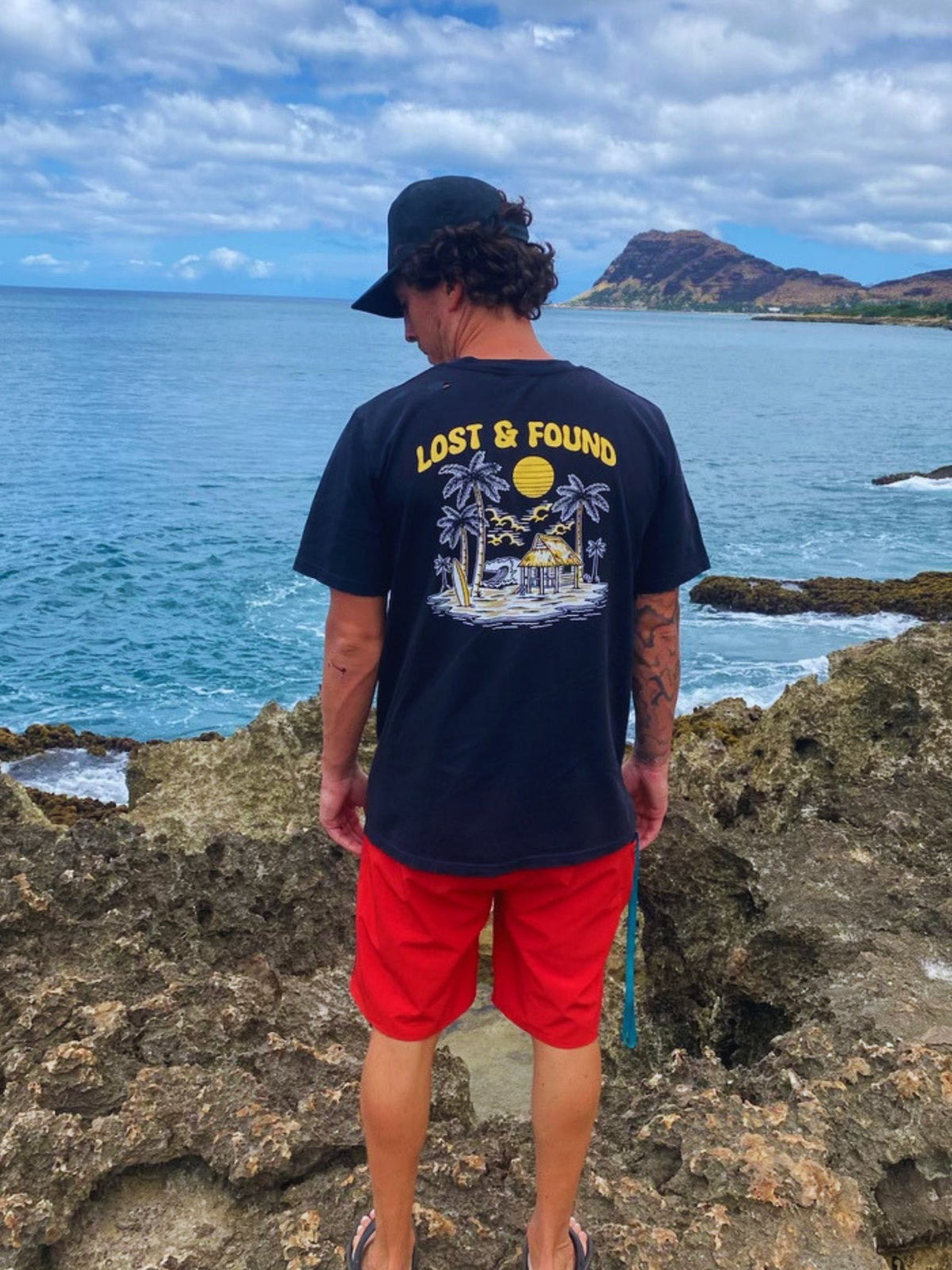 A man stands on a rocky coastline looking out at the ocean, wearing a black "Lost & Found" Seas Get Degrees Tee